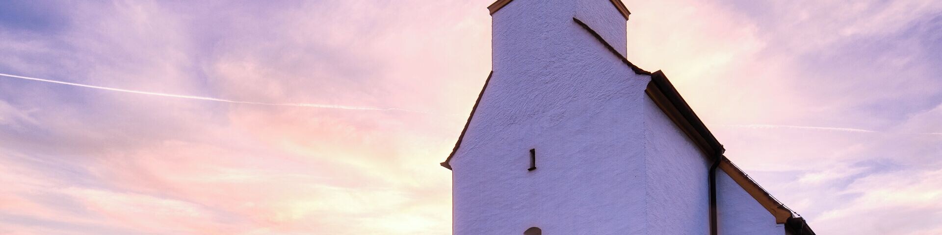 A white church with a black steeple and a clock tower, set against a sunset sky.