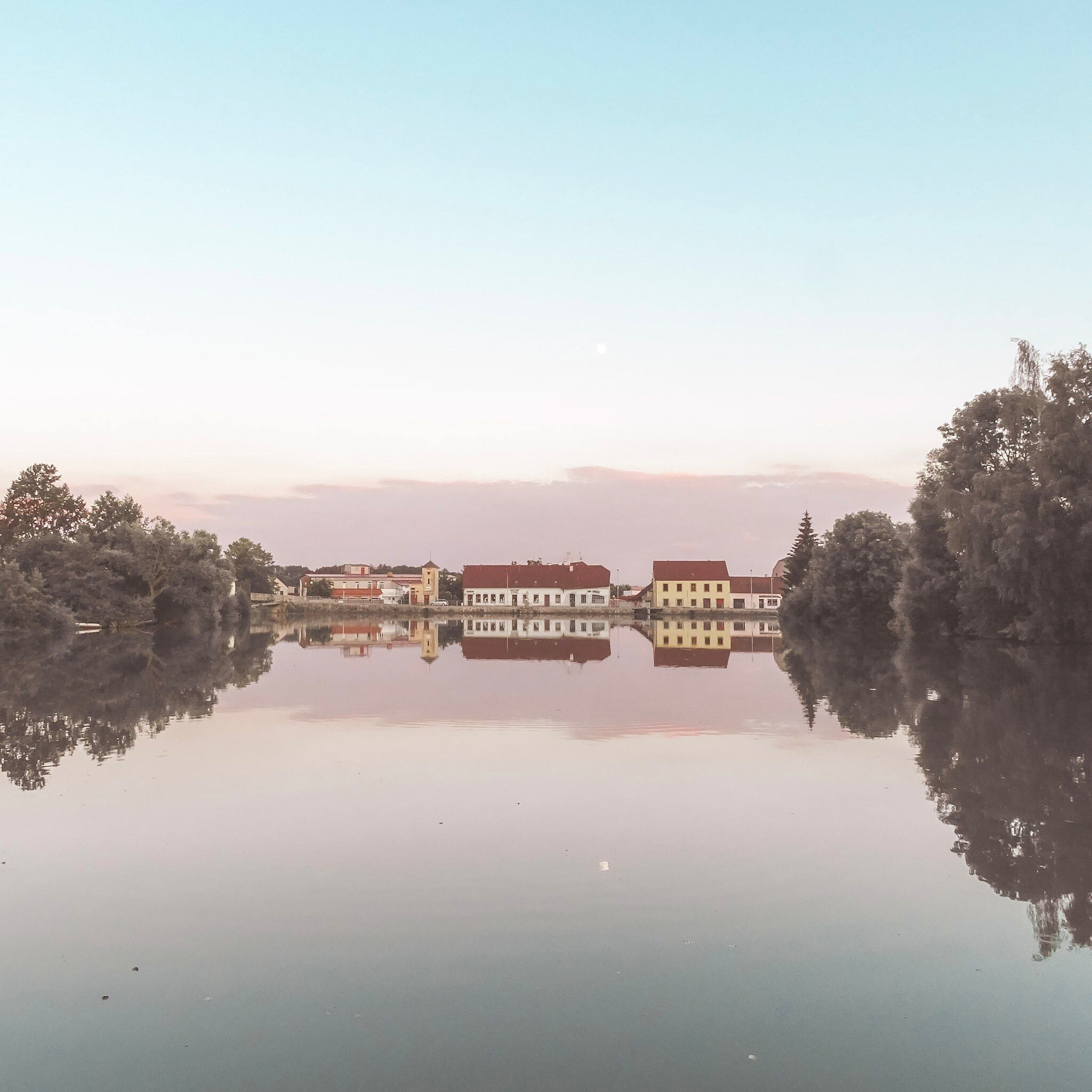 Um lago tranquilo com edifícios refletidos na superfície da água, rodeado por árvores.
