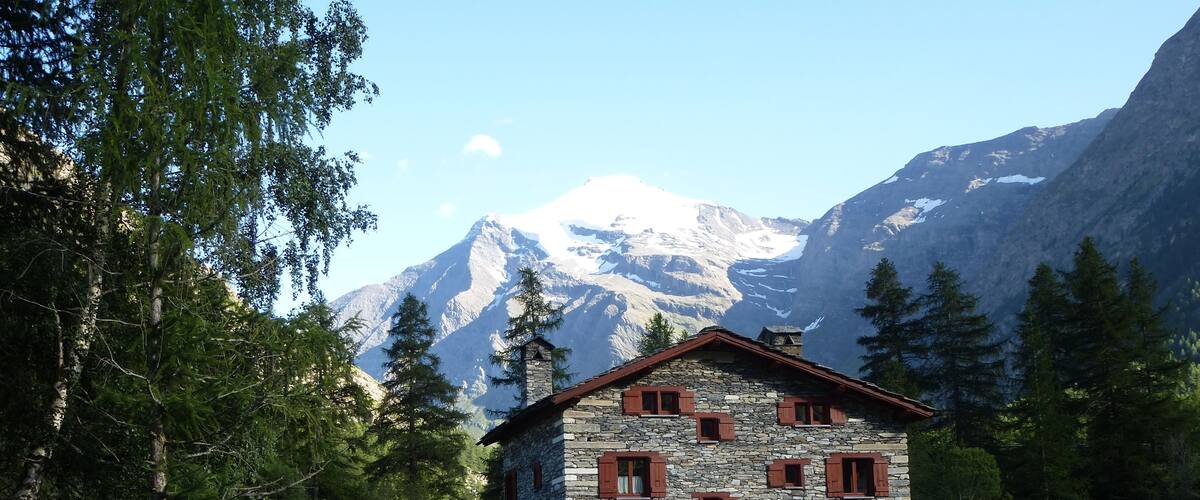A stone house with red shutters, surrounded by lush greenery and a mountain backdrop.