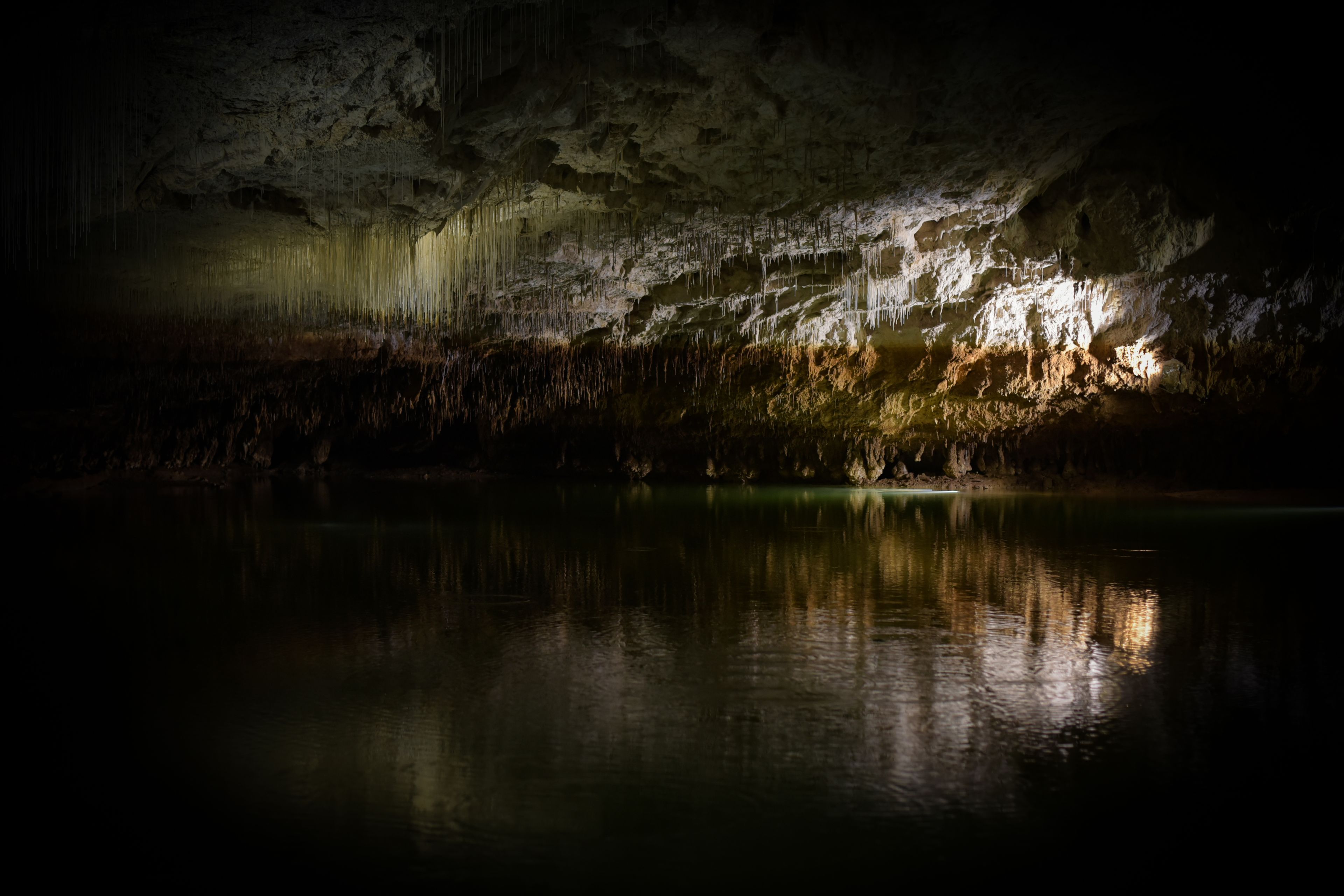 A cave with stalactites and stalagmites, a calm water body reflecting the cave's interior, and a small cave opening.