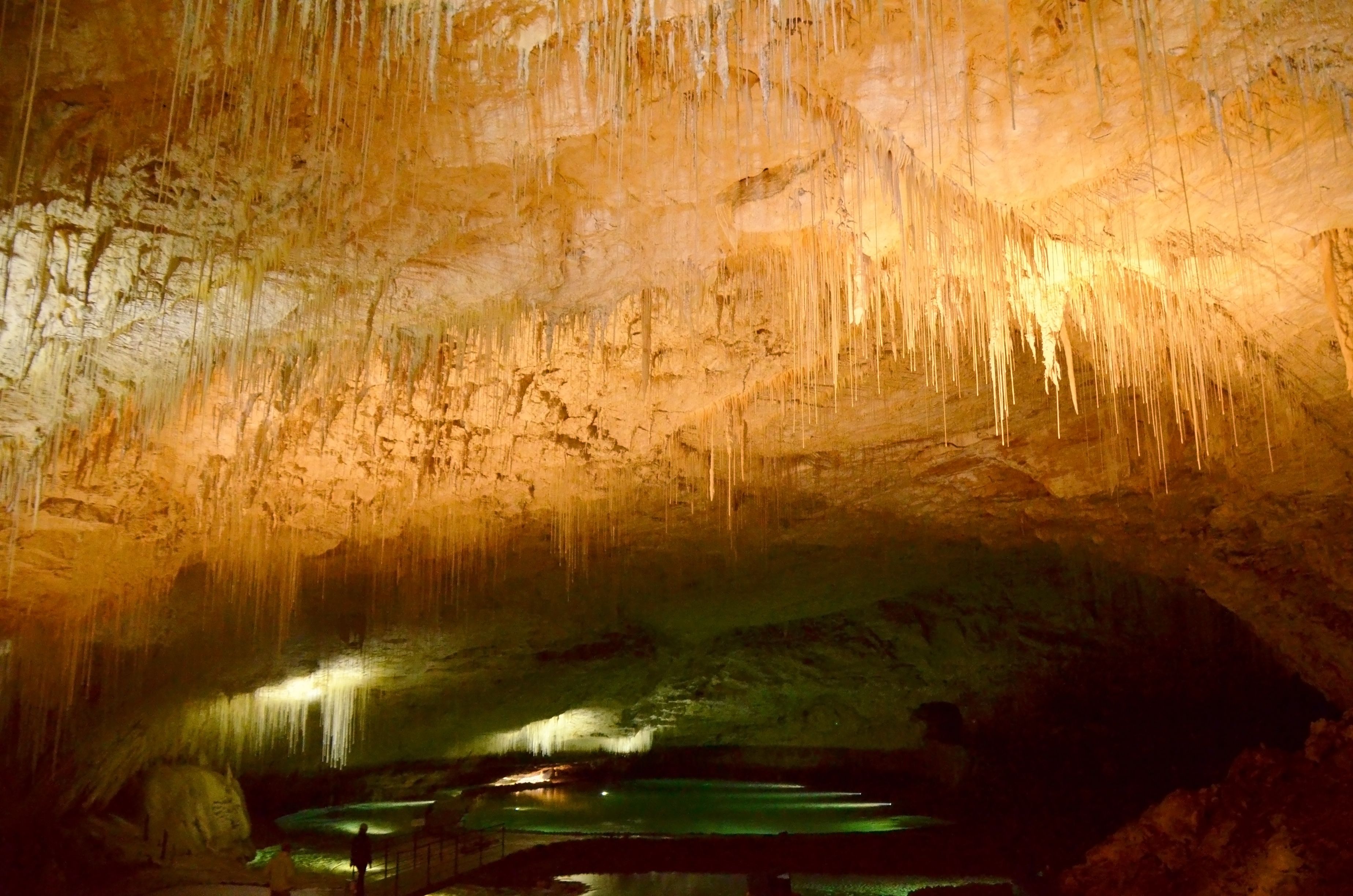 Une grotte ornée de stalactites suspendues au plafond et contenant une mare d’eau au fond.