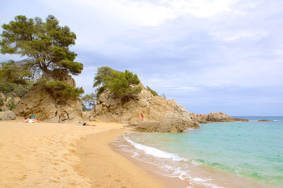 A sandy beach with clear turquoise water, rocky cliffs, and green trees.
