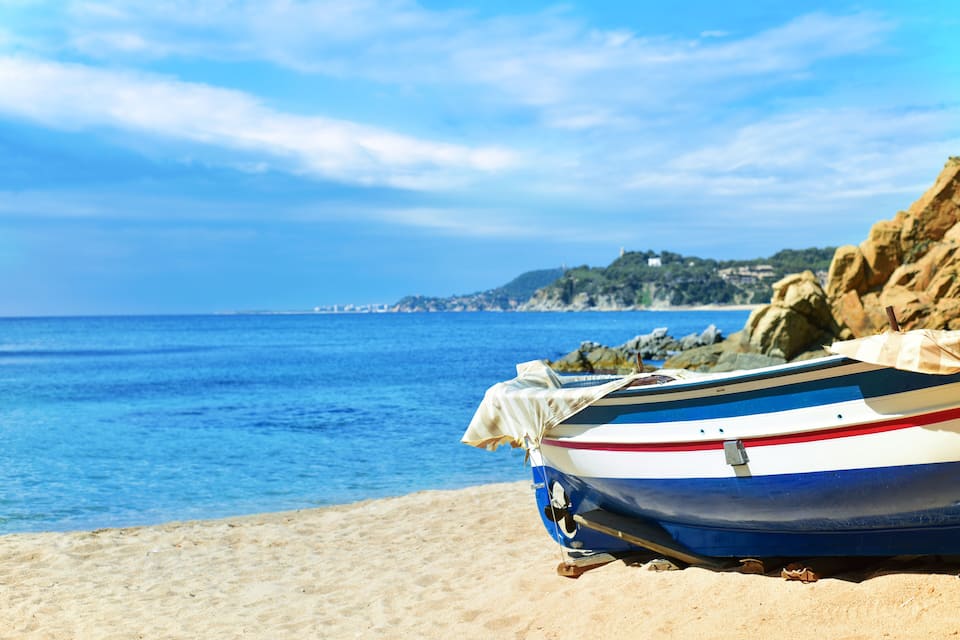 Una piccola barca su una spiaggia sabbiosa, con un mare blu cristallino e scogliere rocciose sullo sfondo.