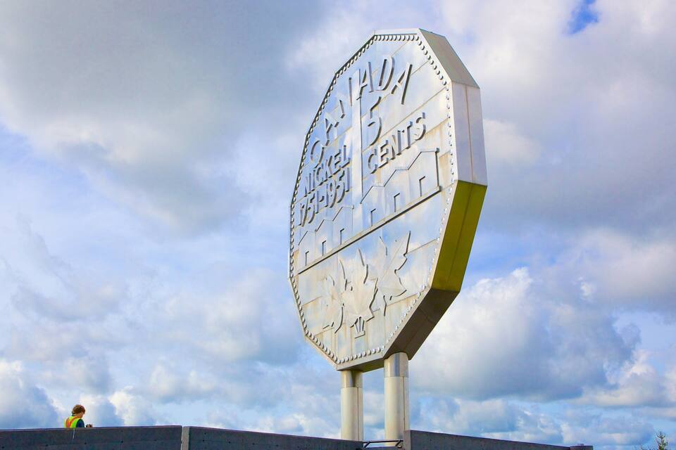 A large, octagonal metal sign with the text "NICKEL CURRENCY CENT" against a partly cloudy sky.