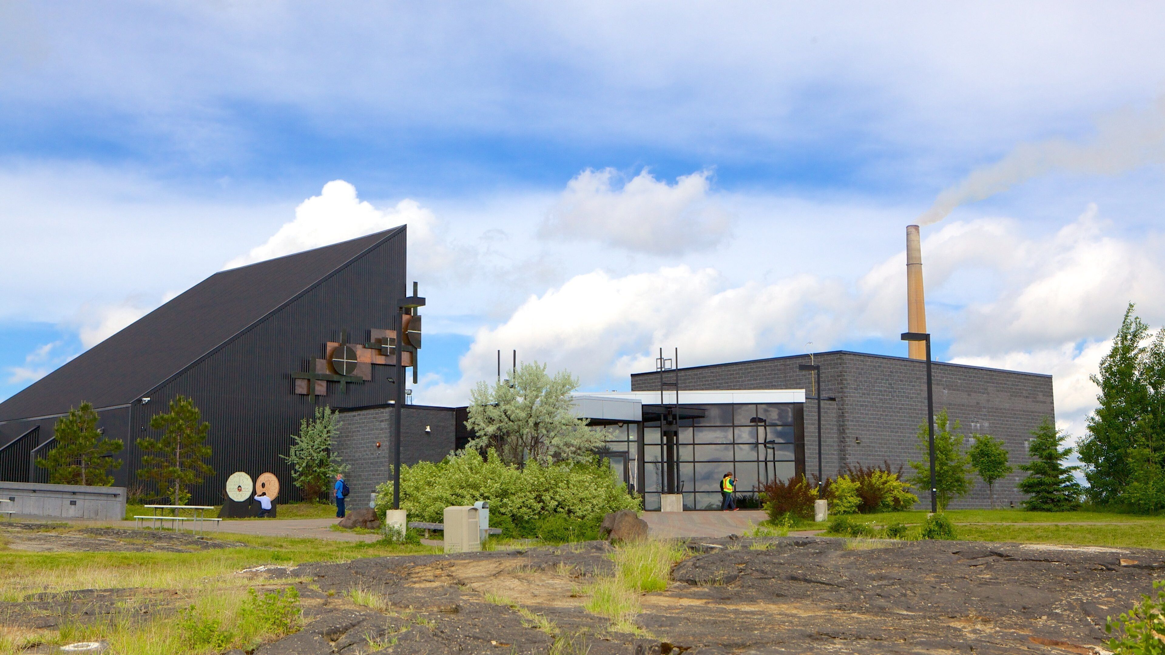 A modern building with a large black roof and a tall chimney emitting smoke.