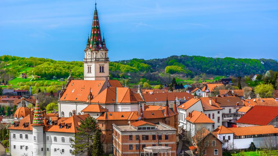 A historic town with a prominent church tower, red-roofed buildings, and lush green hills in the background.