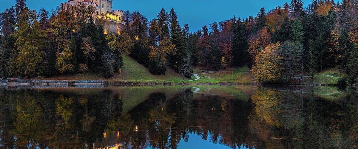 A castle on a hill with autumn trees and a calm lake reflecting the scene.
