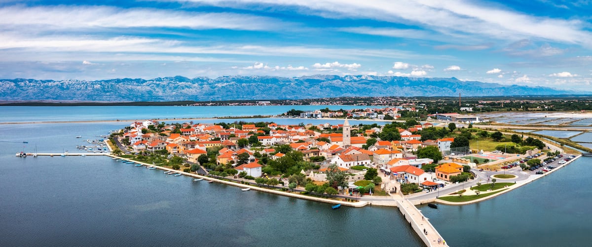 A coastal town with red-roofed buildings, a marina, and mountains in the background.