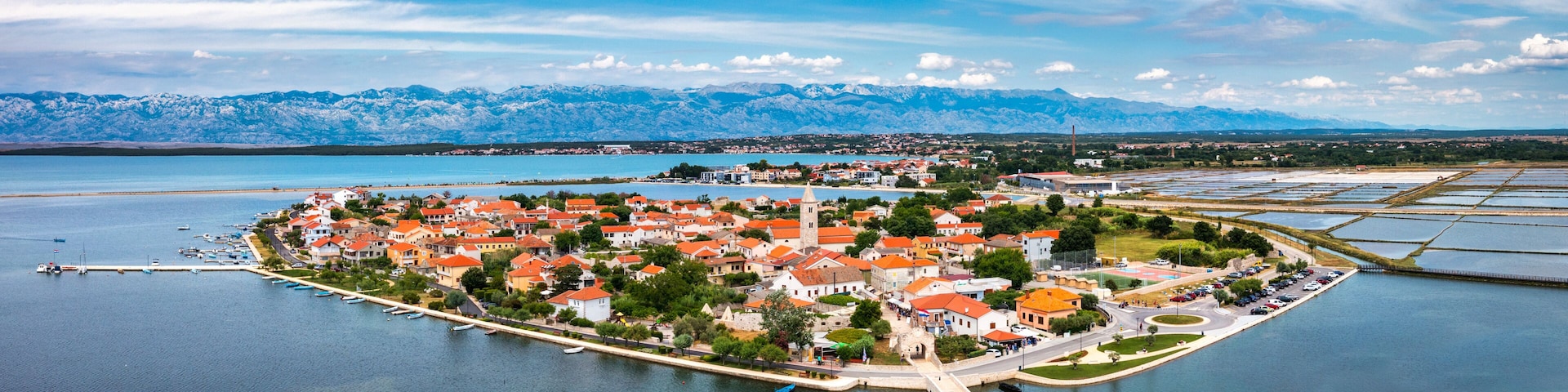 A coastal town with red-roofed buildings, a marina, and mountains in the background.