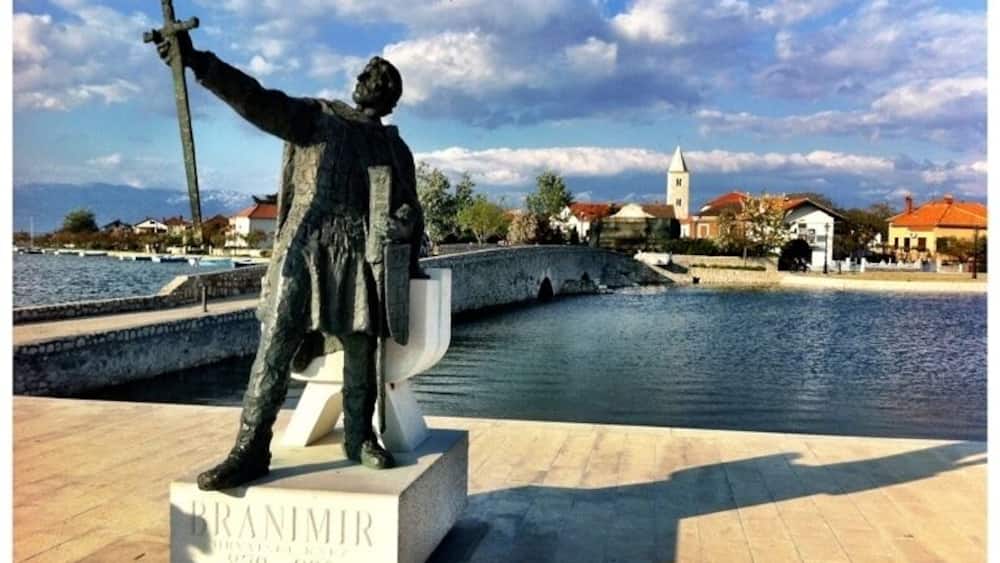 A statue of a man holding a sword, labeled "BRANIMIR," with a body of water and buildings in the background.