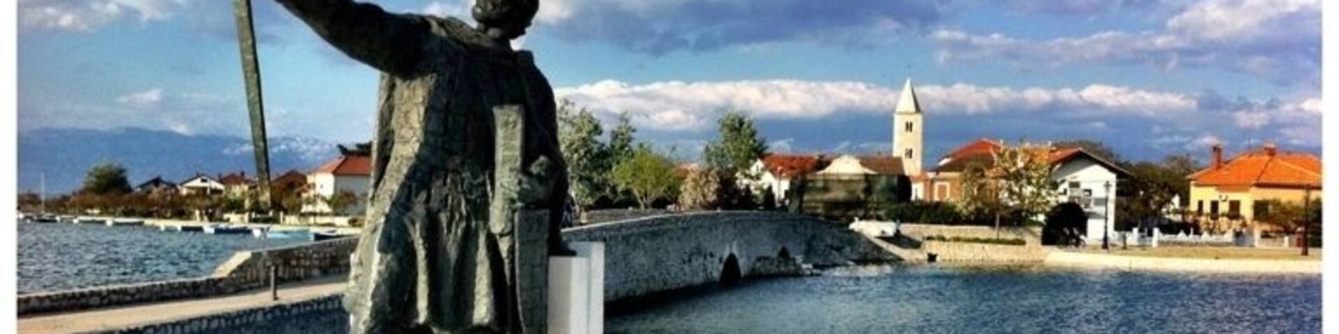 A statue of a man holding a sword, labeled "BRANIMIR," with a body of water and buildings in the background.