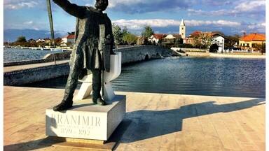 A statue of a man holding a sword, labeled "BRANIMIR," with a body of water and buildings in the background.