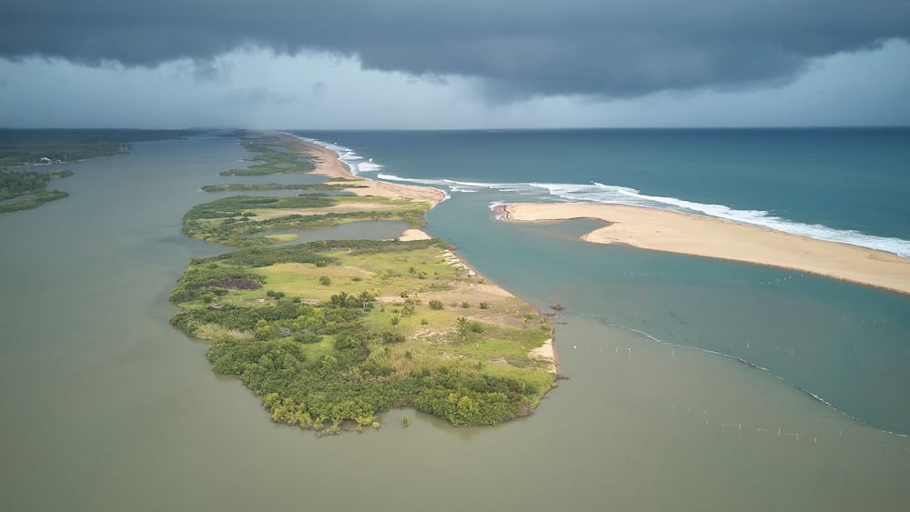 Eine Luftaufnahme einer Küstenlandschaft mit einem weiten Strand, einem Fluss oder einer Lagune und einem stürmischen Himmel.