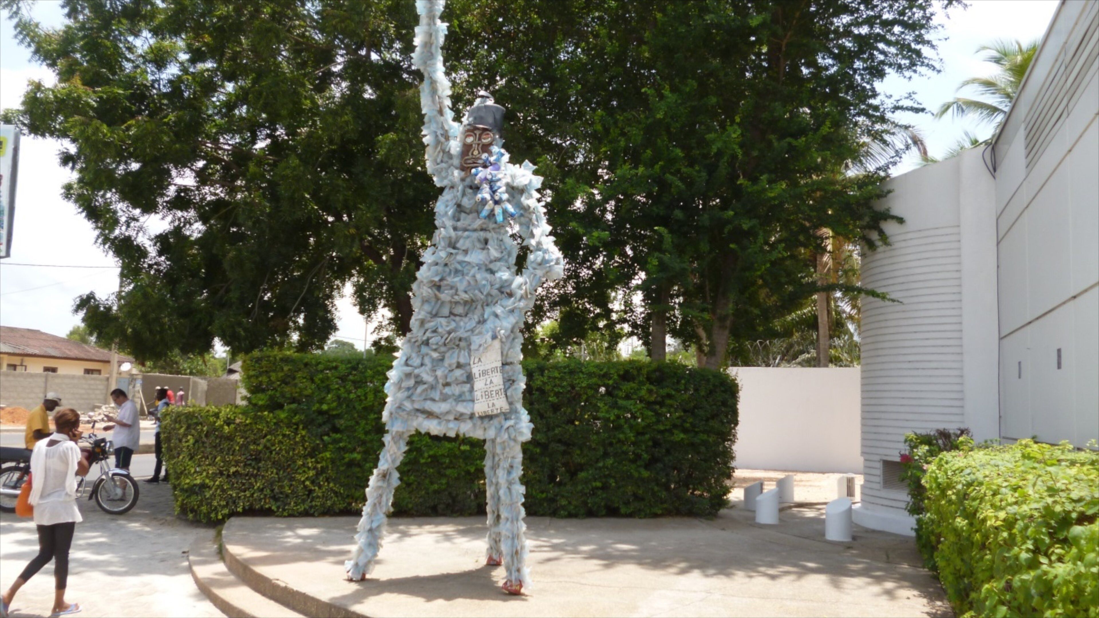 A sculpture of a figure made of white material in an outdoor setting with greenery and buildings in the background.