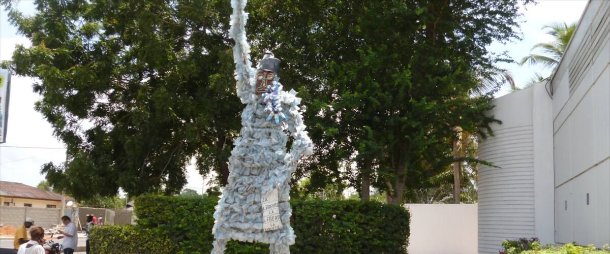A sculpture of a figure made of white material in an outdoor setting with greenery and buildings in the background.