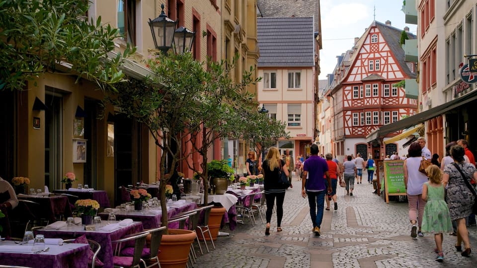 A cobblestone street lined with traditional half-timbered buildings, outdoor dining tables, and pedestrians.