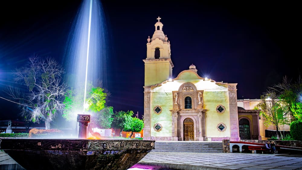 A night scene with a fountain, a church with a bell tower, and a tiled plaza.