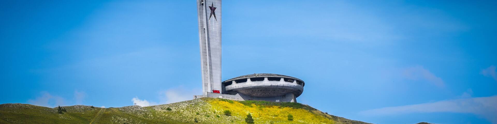 A large, cylindrical monument with a star on top, situated on a grassy hill with a clear blue sky.