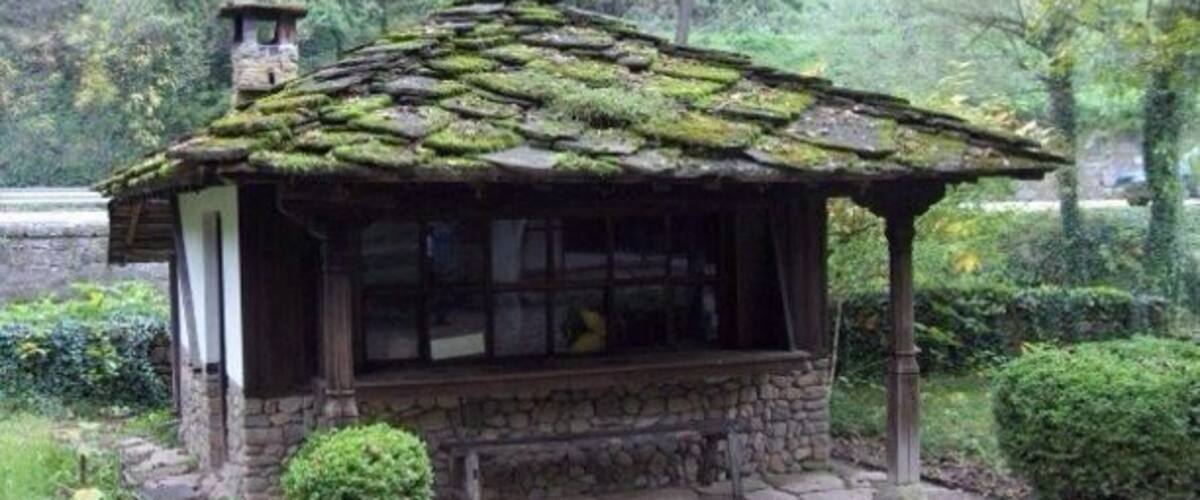A rustic stone gazebo with a moss-covered roof, surrounded by lush greenery and a stone pathway.
