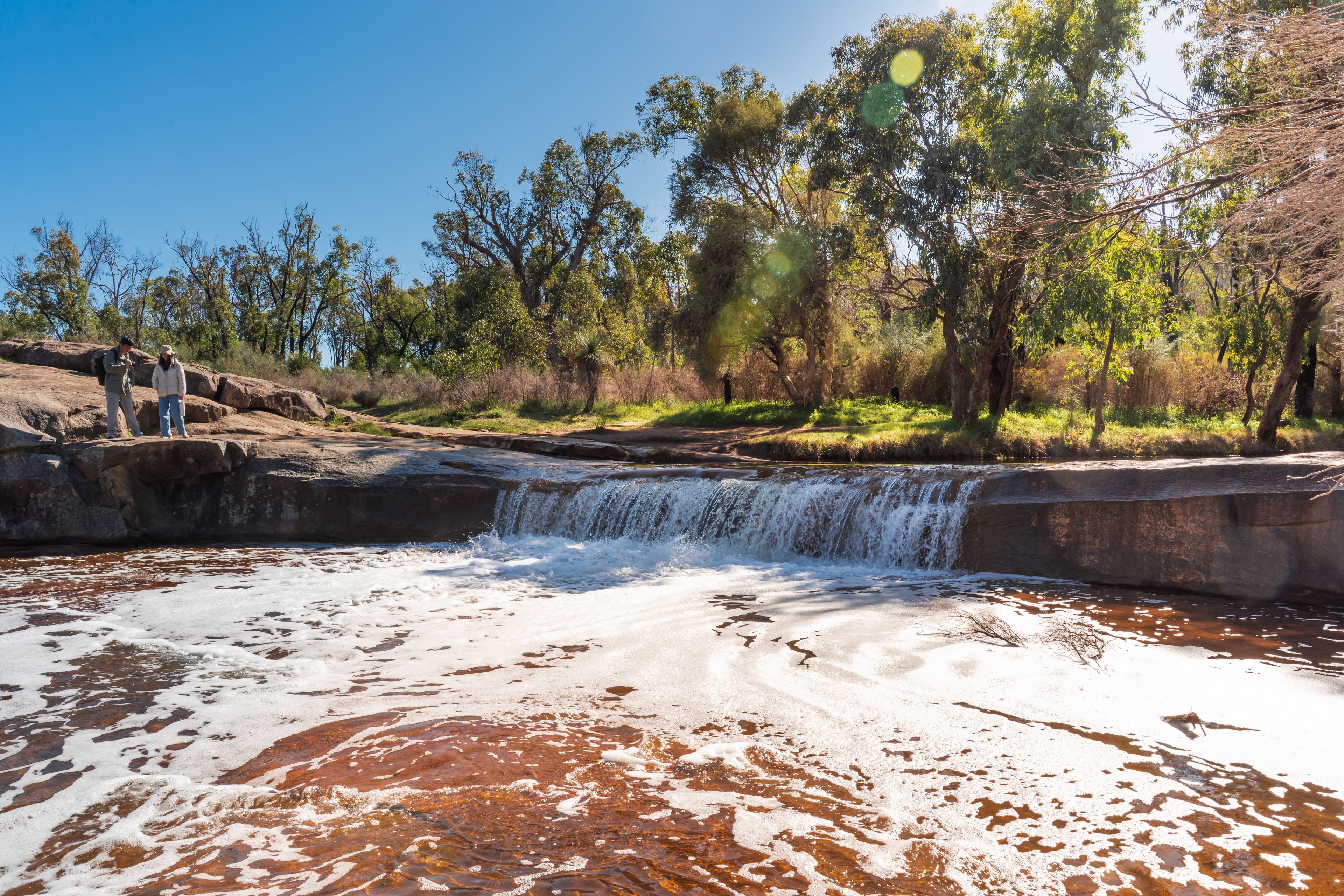 A waterfall flowing over rocks into a shallow pool with a forested area in the background.