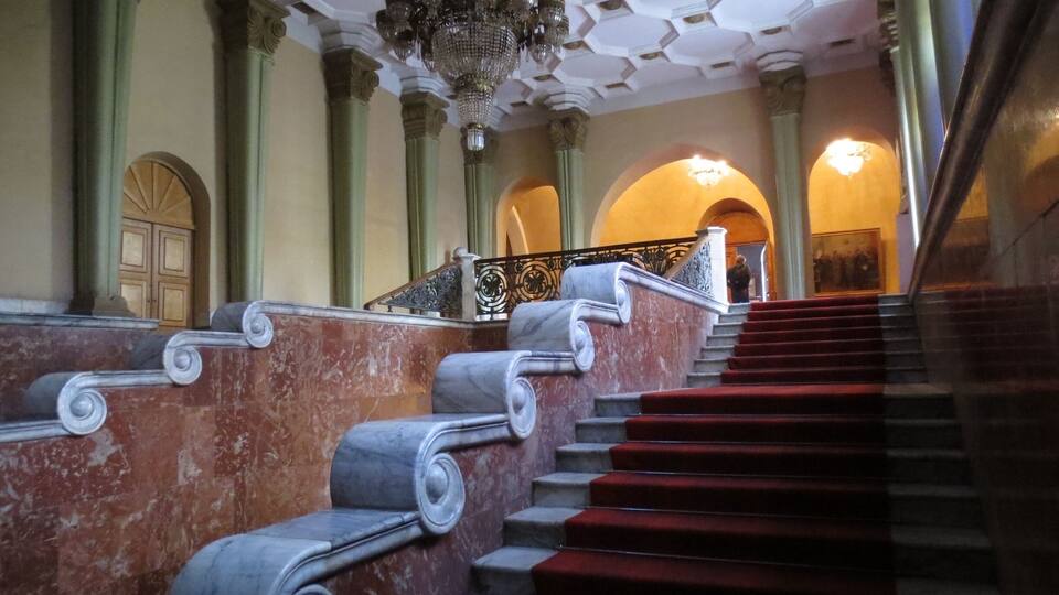 A grand staircase with marble balustrades and red carpeted steps, leading to an arched doorway.