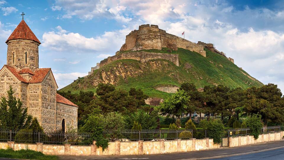 A church with a red roof and a stone tower, and a large hill with a castle on top.