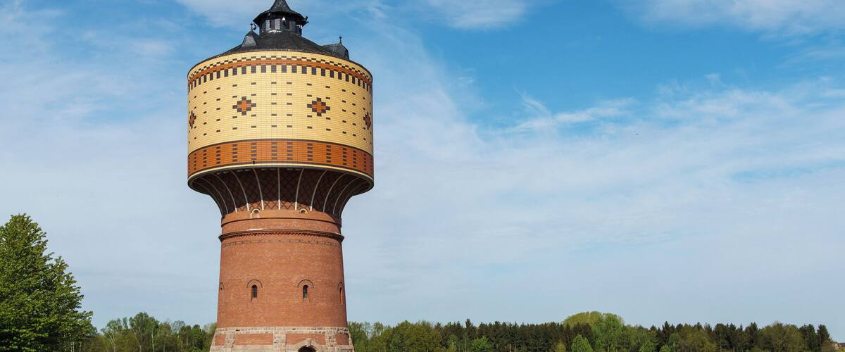 A large water tower with a tiled top and brick base, surrounded by a field and trees.
