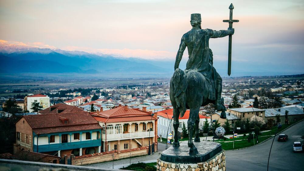 A statue of a figure holding a sword, overlooking a town with red-roofed buildings and mountains in the background.