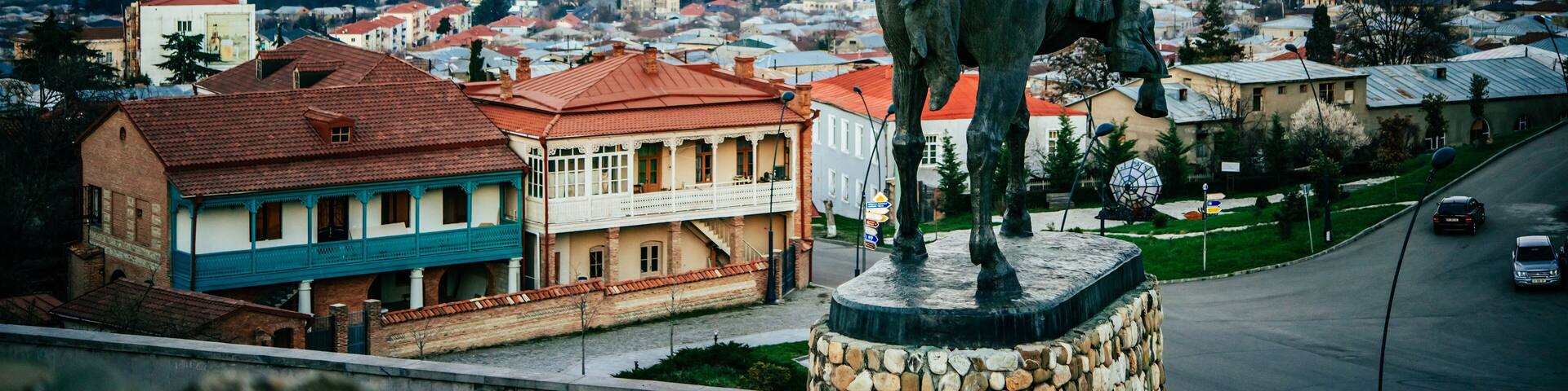 A statue of a figure holding a sword, overlooking a town with red-roofed buildings and mountains in the background.