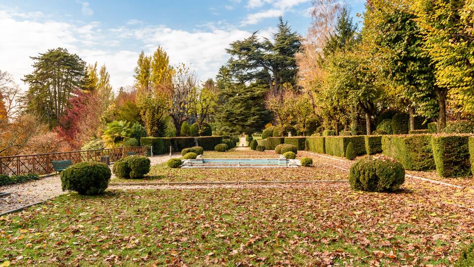 A garden with a fountain, neatly trimmed hedges, and trees with autumn foliage.