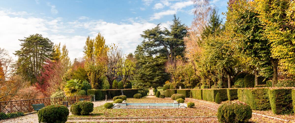 A garden with a fountain, neatly trimmed hedges, and trees with autumn foliage.