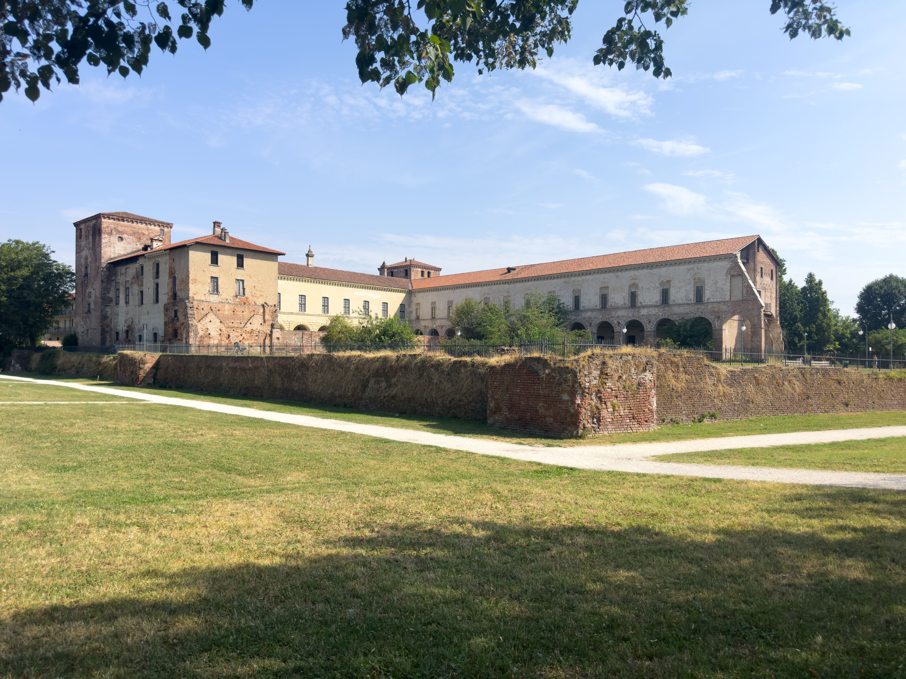 The grand historic Visconti Castle of Melegnano stands majestically behind a lush green park on a sunny day.