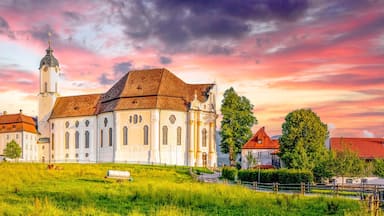 Uma igreja histórica com uma torre imponente, rodeada por vegetação exuberante e um céu poente vibrante.