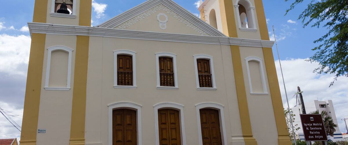 A church with two bell towers, a cross on top, and a sign indicating it is Igreja Matriz N. Senhora Rainha dos Anjos.