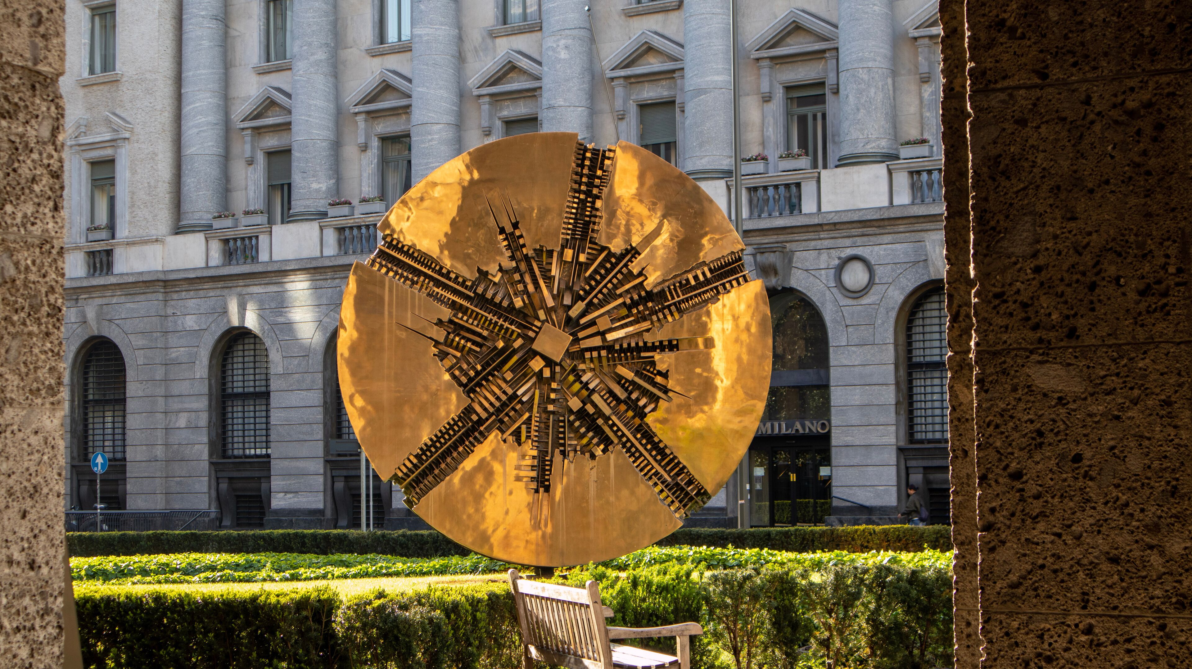A large golden sculpture in front of a historic building with arched windows and a sign reading "MILANO".