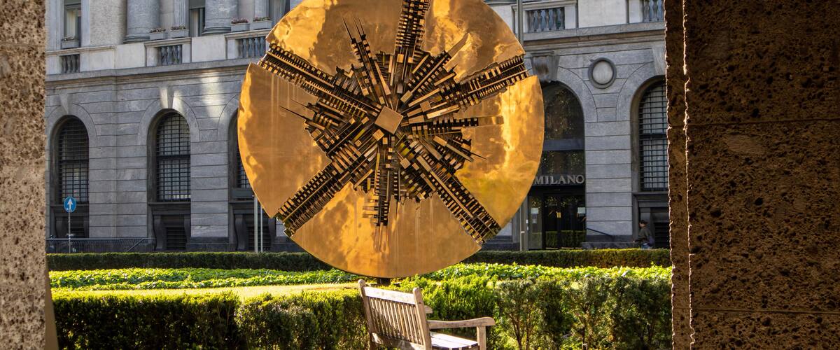 A large golden sculpture in front of a historic building with arched windows and a sign reading "MILANO".