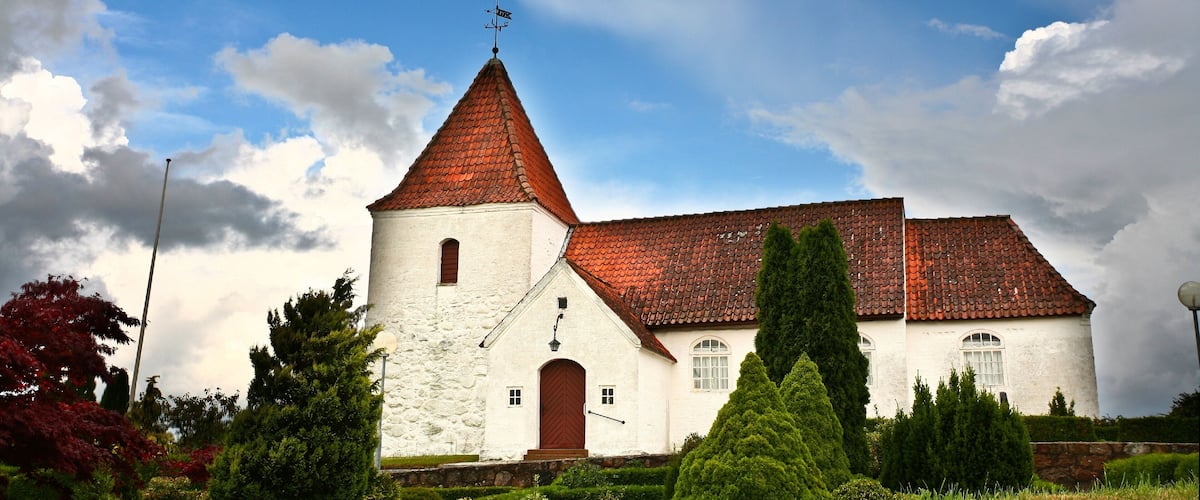 A church with a red-tiled roof and a white facade, surrounded by greenery.