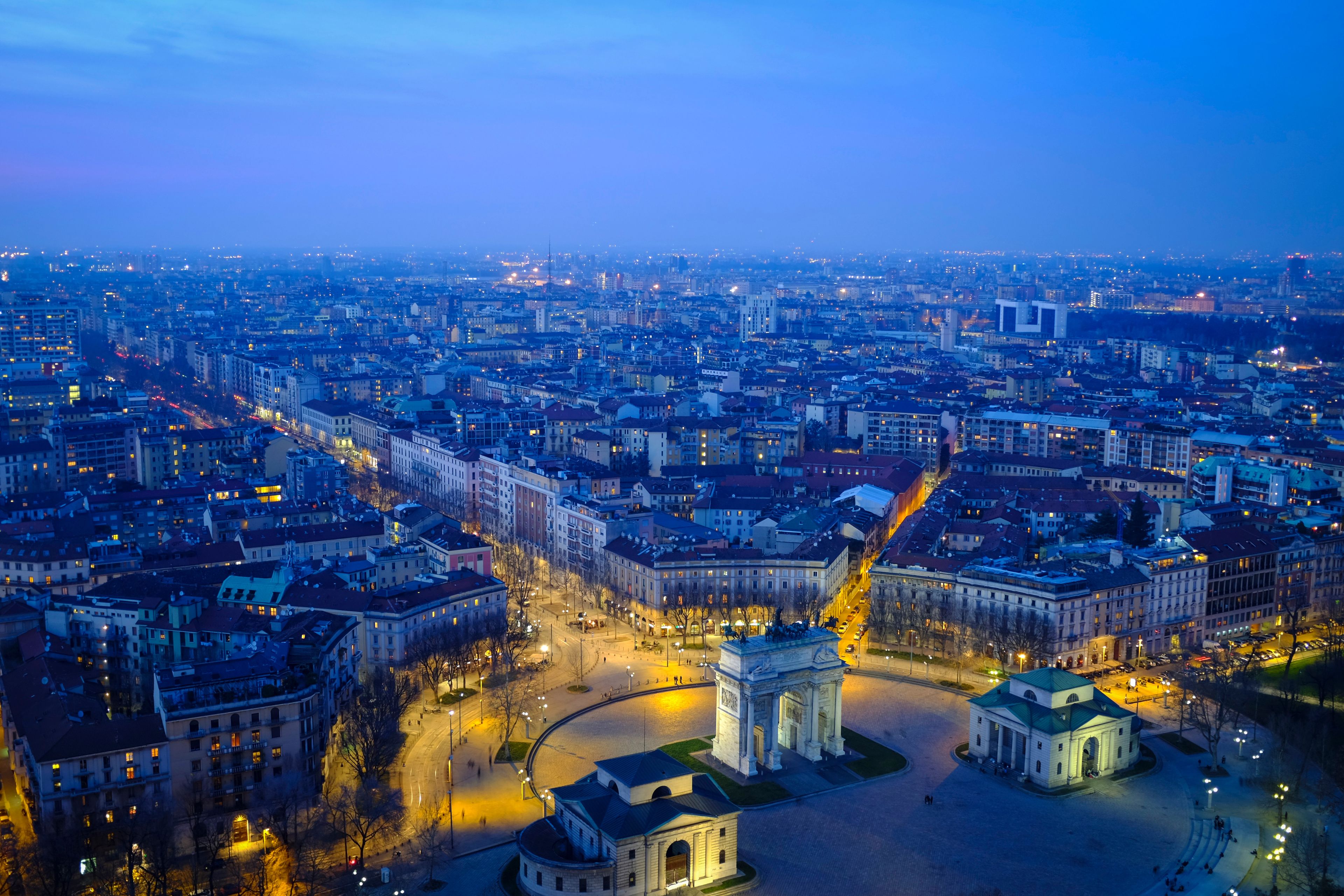 A panoramic view of a cityscape at dusk with illuminated buildings and a prominent monument in the center.