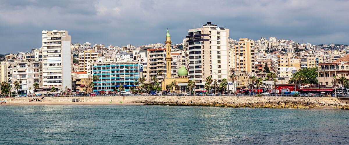 A coastal cityscape with a mix of modern and traditional buildings, a prominent mosque, and a rocky shoreline.