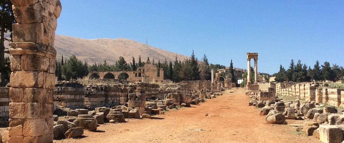 Ancient ruins with stone columns and arches, a dirt path, and a mountain in the background.