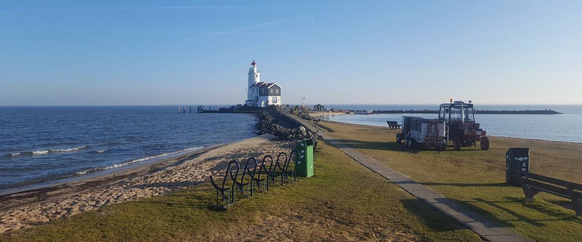 Un faro sobre un promontorio rocoso, una playa con valla y un tractor estacionado cerca del agua.
