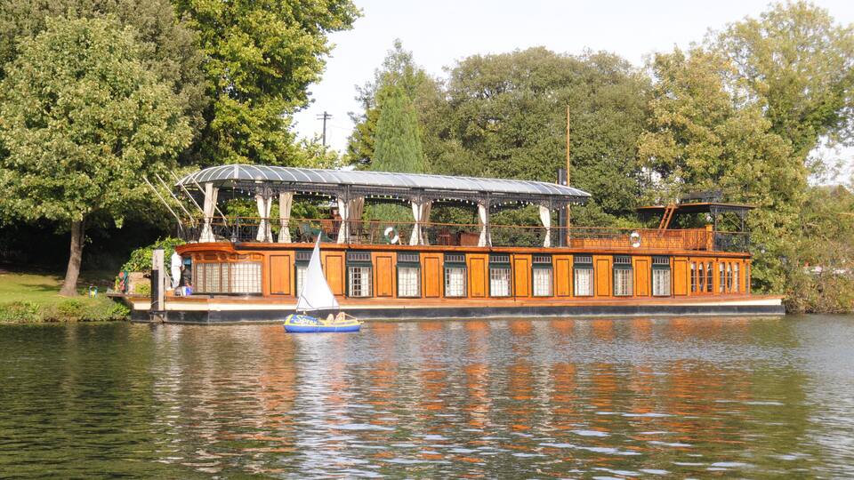 A houseboat with a covered deck and orange exterior, docked on a calm lake surrounded by trees.