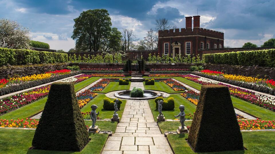 A formal garden with a central fountain, symmetrical flower beds, and statues.