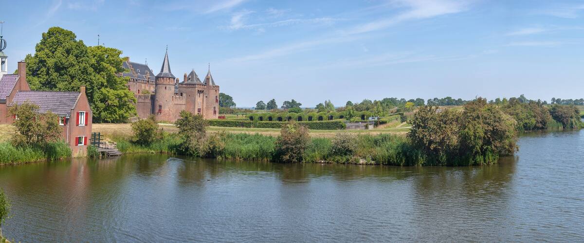 A historic castle by a calm river, surrounded by lush greenery and wind turbines in the distance.