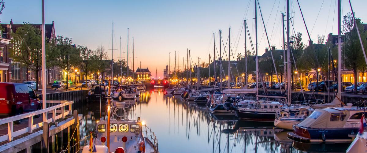 A marina with boats docked, a red van parked on the left, and a calm water reflecting the surroundings.