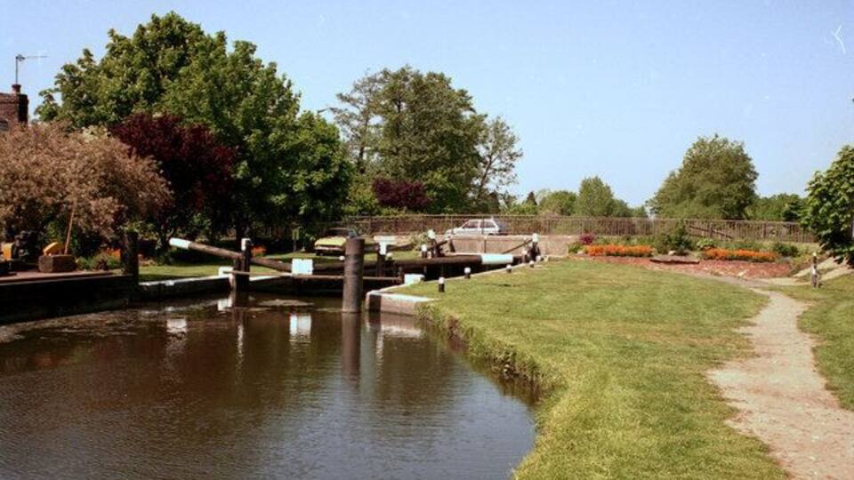 A calm river with a grassy bank, a wooden bridge, and trees in the background.
