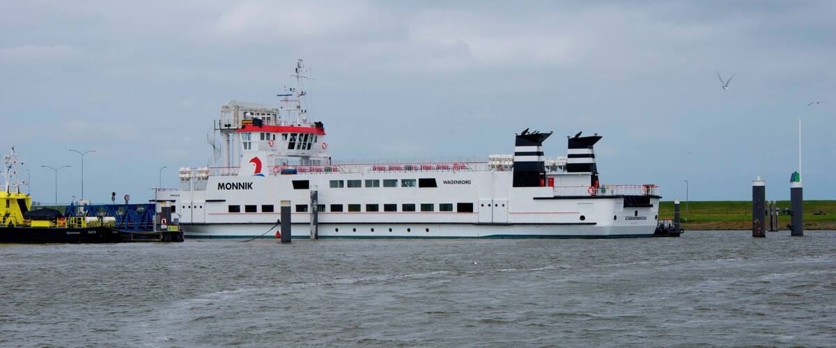 A large white ferry with 'MONNIK' and 'WAGENBORG' written on it, docked at a harbor with calm water and a cloudy sky.