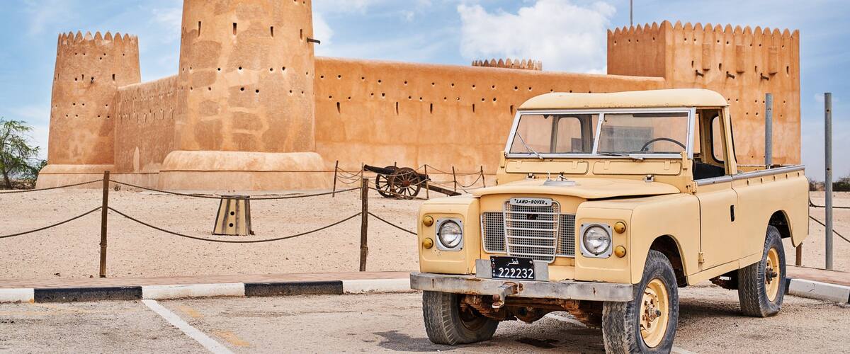 A beige Land Rover in front of a large, ancient fort with multiple towers and a flag.