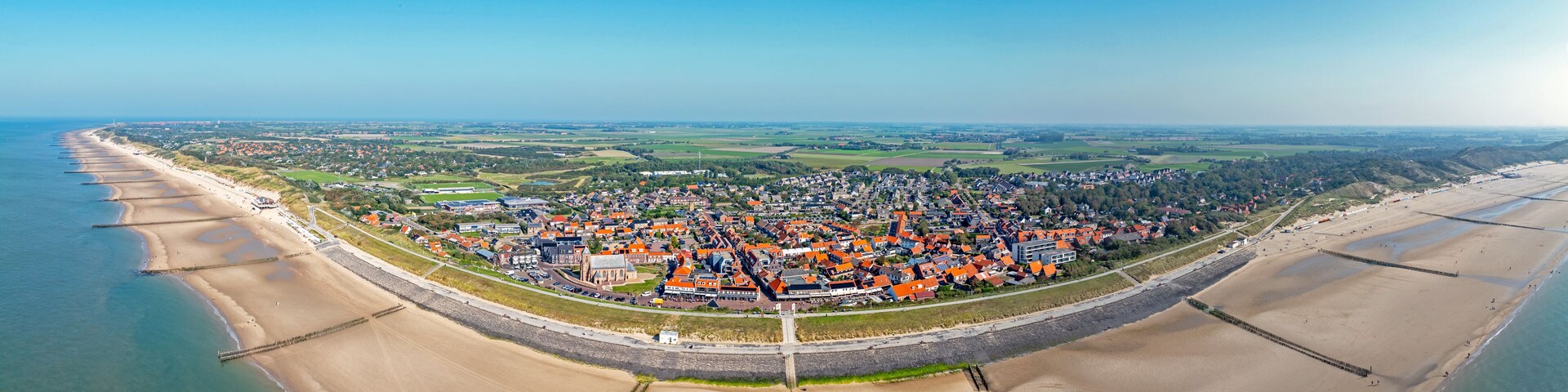 Vista aérea de un pueblo costero con playa, un muro fortificado y campos circundantes.