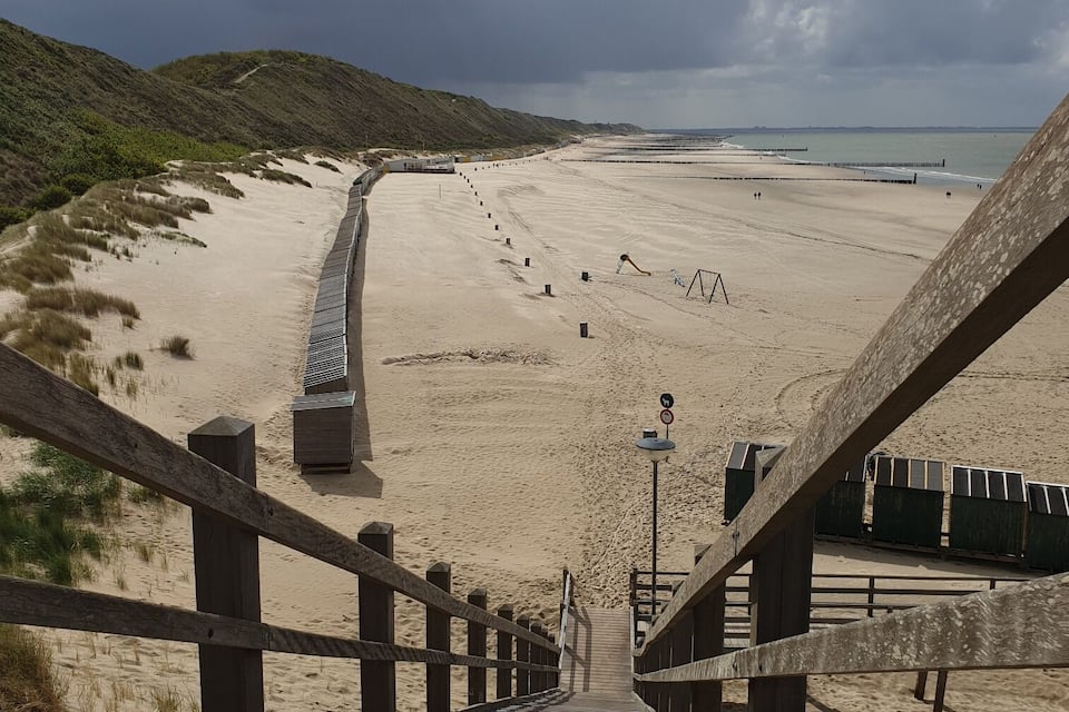 Ein hölzerner Steg führt zu einem Sandstrand mit Dünen und bewölktem Himmel.